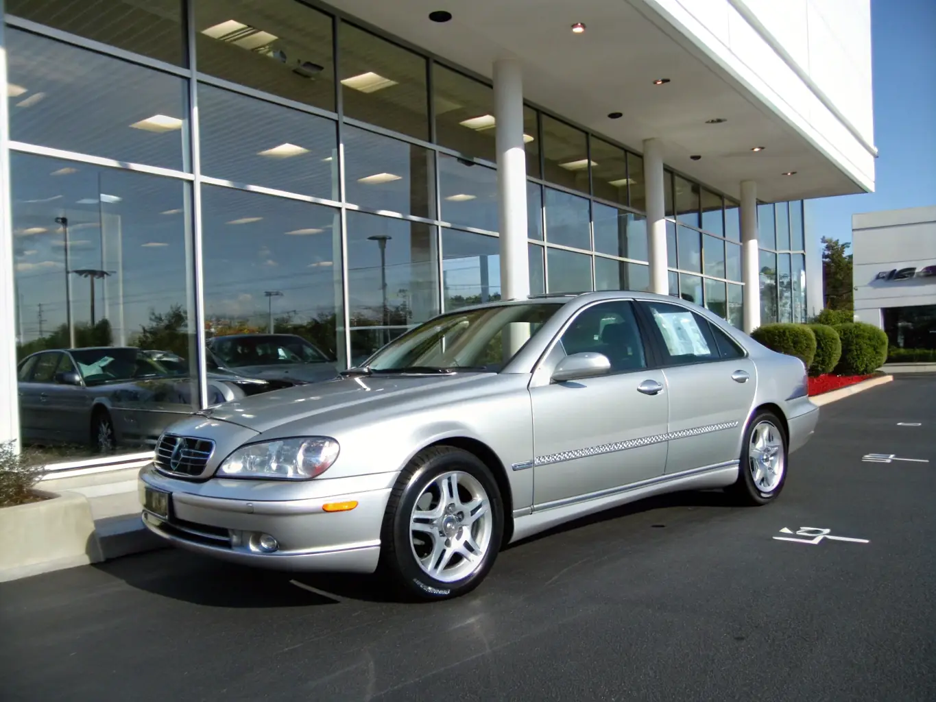 A modern car carrier truck is shown delivering a sleek sedan to a car dealership, with the dealership's logo visible in the background, under a sunny sky.